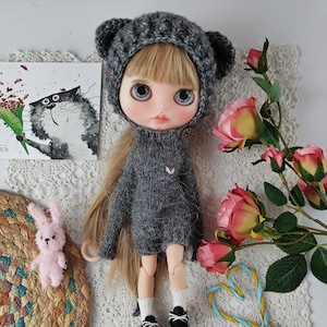 Puede incluir: Una muñeca de punto gris con un gorro de punto gris y grandes ojos azules. La muñeca lleva un suéter de punto gris y tiene el pelo largo y rubio. La muñeca está sentada sobre una superficie blanca con un patrón floral.