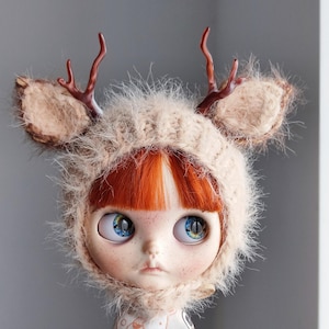 Pode incluir: Uma boneca usando um chapéu de tricô bege com orelhas e chifres de veado marrons e peludos. O chapéu tem uma textura felpuda e a boneca tem olhos azuis e cabelo ruivo.