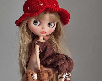 May include: A doll with long blonde hair, wearing a red strawberry-shaped hat with white heart accents. The doll is dressed in a brown jumpsuit with floral cuffs and red shoes. A small brown teddy bear is held in the doll's hand.