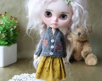 Puede incluir: Una muñeca con el pelo largo, blanco y rizado que lleva un cárdigan de punto gris con detalles rojos y naranjas y una falda amarilla con estampado de pájaros.