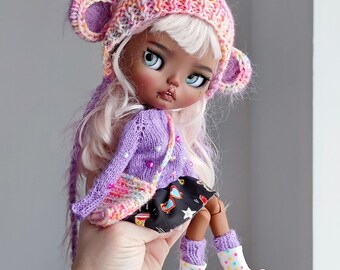 May include: A doll with pink hair and brown skin wearing a purple sweater, a black and white skirt, and a pink and white knitted hat with bear ears and a gold unicorn horn. The doll is wearing white rain boots with colorful polka dots.