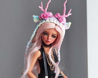 Peut inclure: Une poupée aux longs cheveux roses portant un haut noir et un chapeau crocheté avec des bois et des roses roses. La poupée tient un appareil photo noir avec une sangle noire et une chaîne dorée.