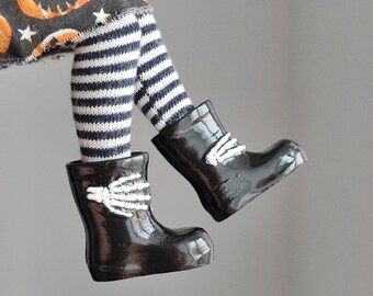 May include: Black rain boots with white skeleton hand designs. The boots are worn by a doll with striped tights and a pumpkin-patterned skirt.