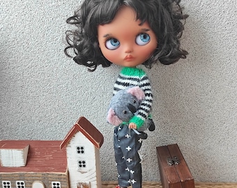 Puede incluir: Una muñeca con el pelo largo negro y rizado, ojos azules y piel morena lleva un jersey a rayas verdes y negras y pantalones negros con un patrón de cruz blanca. La muñeca sostiene un koala de peluche gris.