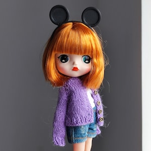 May include: A doll with orange hair, wearing a purple knitted cardigan, denim shorts, and a black headband with mouse ears. The doll has large eyes and red lipstick. The doll is a collectible item.