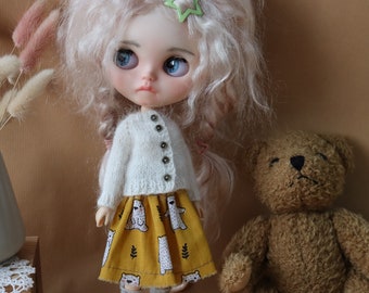 Puede incluir: Un cárdigan blanco de punto con cierre de botones, usado por una muñeca con cabello rubio y ojos azules. La muñeca lleva una falda amarilla con estampado de osos polares blancos. Un oso de peluche marrón está sentado en el fondo.