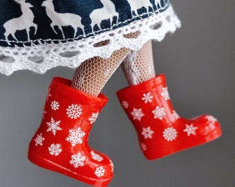 May include: Red rain boots with white snowflake designs. The boots are worn by a doll with white lace trim on a blue and white reindeer patterned skirt.
