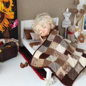 Puede incluir: Una muñeca con cabello rubio y ojos cerrados está durmiendo en una cama blanca con una colcha de retazos marrones y blancos. La cama está en un entorno de casa de muñecas con una maleta marrón y un oso de peluche.