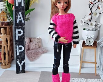 Puede incluir: Una muñeca con un suéter a rayas rosa y negro de manga larga y un conjunto de pantalones de cuero negro. La muñeca tiene el pelo largo y rubio y lleva zapatos amarillos. La muñeca está de pie frente a un sofá blanco y una torre de bloques de madera que deletrea "HAPPY".