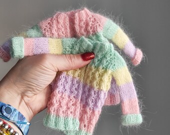 May include: Two pastel-colored knitted sweaters. The sweaters are held in a hand and feature a striped design in pink, green, yellow, and purple. The sleeves and body have different textures. The sweaters are small, possibly for a doll or a baby.