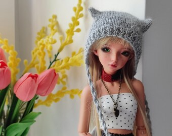 Peut inclure: Une poupée porte un chapeau gris en tricot avec des oreilles de chat et de longues tresses. La poupée a les cheveux blonds, les yeux verts et porte un haut tube blanc à pois noirs, un ras du cou rouge et noir et un collier pendentif. Des tulipes roses et du mimosa jaune en arrière-plan.