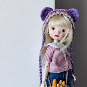 Puede incluir: Una muñeca con cabello rubio que lleva un gorro de punto morado y blanco con orejas de oso y una camisa a rayas rosa y blanca. La muñeca sostiene una cesta de pan.