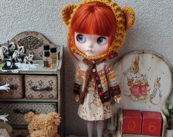 Puede incluir: Una muñeca con cabello rojo y ojos azules que lleva un gorro de oso amarillo, un suéter marrón y naranja y un vestido floral. La muñeca está frente a un pequeño armario de madera y un calendario decorativo.