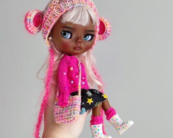May include: A doll with dark brown skin, white hair, and blue eyes is wearing a pink knitted hat with bear ears, a pink sweater, a black and white skirt, and pink and white rain boots. The doll is holding a pink and white crocheted bag.