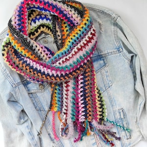 Puede incluir: Una bufanda de crochet colorida con una variedad de colores, incluyendo rosa, azul, verde y amarillo. La bufanda está drapeada sobre una chaqueta vaquera.