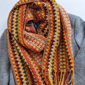 May include: A crocheted scarf in a granny square design. The scarf displays a warm colour scheme of orange, yellow, brown, and tan. It has a fringed edge and is draped over a grey coat.