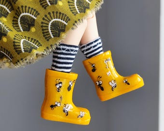 May include: Yellow rain boots with a black and white striped pattern on the top of the boot. The boots have a cartoon design of insects on them.