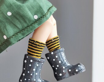 May include: A pair of black rain boots with white polka dots. The boots are worn by a doll with yellow and black striped socks.