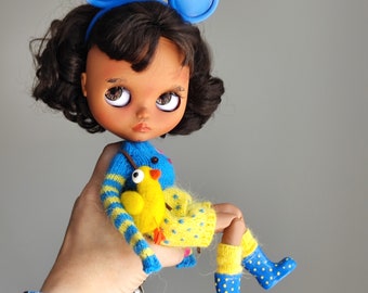 May include: A doll with brown hair and blue eyes wearing a blue and yellow knitted sweater and blue rain boots. The doll is holding a yellow chick toy.
