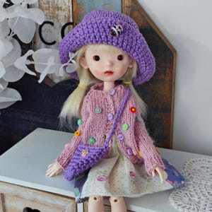 Può includere: Una bambola con un cappello all'uncinetto viola con un ornamento a coccinella, un cardigan rosa decorato con accenti floreali colorati e una borsetta viola abbinata. La bambola ha i capelli biondi e indossa un vestito con stampa floreale.