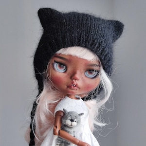 Op de afbeelding: Een pop met een zwarte kattenoormuts, wit haar en een witte jurk. De pop heeft een lichte teint met roze wangen en blauwe ogen. Ze houdt een kleine grijze stoffen wolf vast.