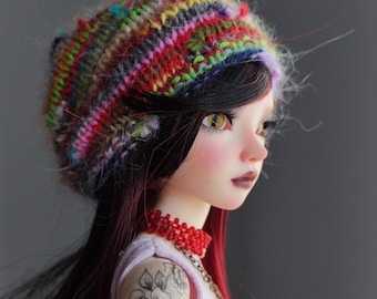 Peut inclure: Une poupée aux longs cheveux rouge foncé portant un bonnet tricoté coloré avec une texture moelleuse. La poupée a un teint clair et des yeux bruns. Elle porte une chemise rose sans manches et un collier de perles rouges.