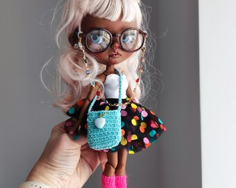 May include: A fashion doll with light pink hair, wearing large round glasses, a white top, and a black skirt with colorful polka dots. The doll has a small blue crocheted purse and is wearing pink leg warmers and blue boots.