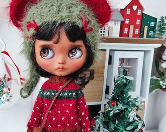 May include: A doll wearing a red and green sweater, green floral skirt, and a green hat with red ear-shaped accents. The doll has large, expressive eyes and a small brown purse. A miniature Christmas tree and decorative houses are in the background.