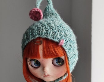 Puede incluir: Una muñeca con cabello rojo y ojos azules que lleva un gorro de punto azul claro con un pompón rosa y una bufanda a juego. El gorro tiene forma puntiaguda y un pequeño acento rosa. La cara de la muñeca tiene pecas y una expresión seria.
