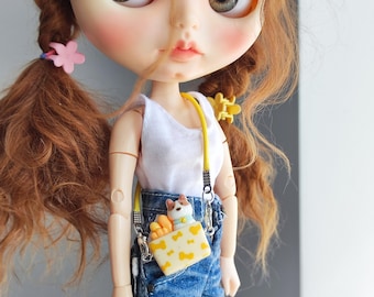 May include: A doll with auburn hair styled in pigtails, wearing a white tank top and denim jeans. A yellow crossbody bag with a cat and bone pattern is attached to the doll. The doll has large eyes and is wearing a small pink flower hair clip.