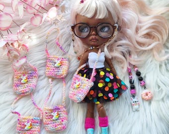 May include: A doll with light pink hair and glasses, wearing a white top and a black skirt with colorful polka dots. Several small, colorful crochet purses with unicorn appliques are displayed. The doll is wearing pink socks and blue boots.