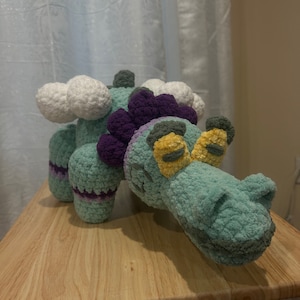May include: A handmade crochet dinosaur in teal, purple, yellow, and white. The plush toy has a long neck, a detailed head with yellow eyes, and cloud-like shapes on its back. It is displayed on a wooden table.
