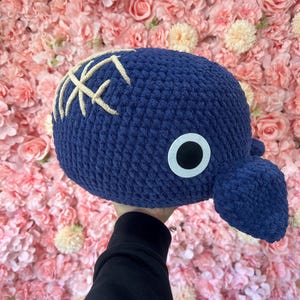 Könnte beinhalten: Ein marineblaues gehäkeltes Wal-Plüschtier mit einem weißen Auge und beigen Kreuzstichdetails. Das Plüschtier hat einen abgerundeten Körper und einen kleinen Schwanz. Der Hintergrund ist eine Wand aus rosa und cremefarbenen Blumen.