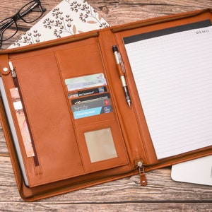 May include: A brown leather portfolio with a zipper closure, a pen holder, and multiple card slots. The portfolio is open to reveal a lined notepad with the word "MEMO" printed at the top.