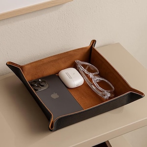 May include: A black and brown leather tray with a phone, wireless earbuds, and clear glasses. The tray is on a beige surface. The tray has a square shape with raised edges.