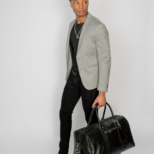 May include: A man wearing a gray blazer, black t-shirt, black jeans, and white sneakers is carrying a black leather duffel bag.