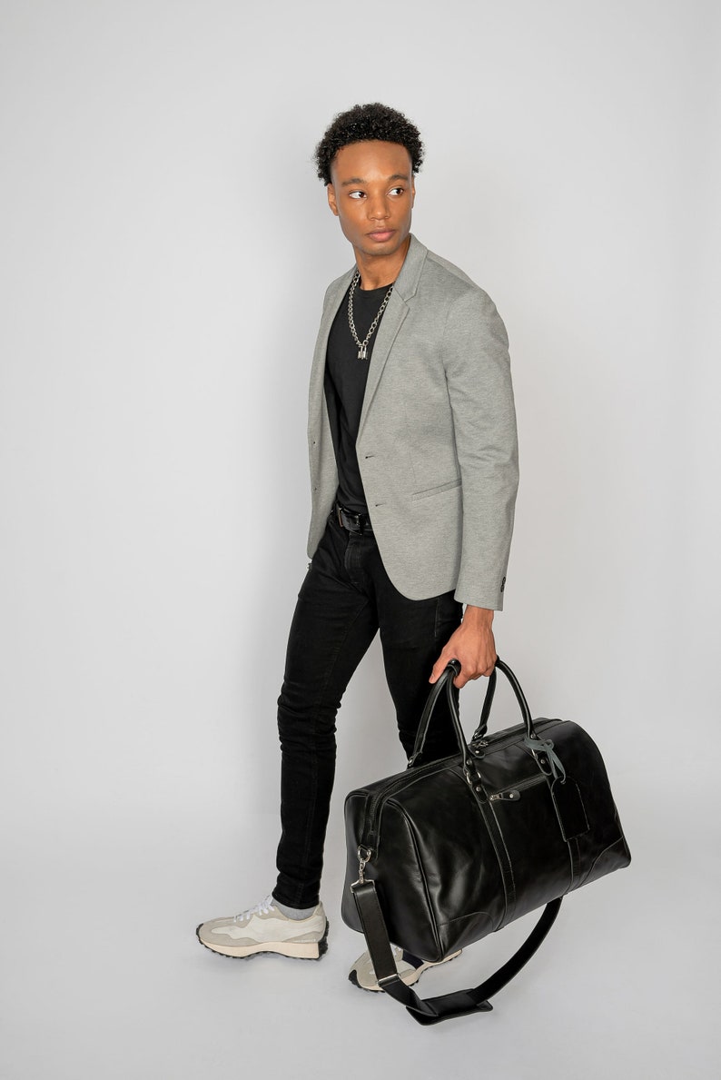 May include: A man wearing a gray blazer, black t-shirt, black jeans, and white sneakers is carrying a black leather duffel bag.