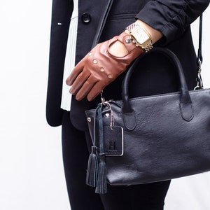 Puede incluir: Un bolso de mano de cuero negro con asa superior y bandolera. El bolso tiene un peque&ntilde;o charm plateado con un borla adjunto. El bolso est&aacute; siendo sostenido por una persona que lleva una chaqueta negra y guantes de cuero marr&oacute;n.
