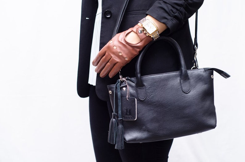Puede incluir: Un bolso de mano de cuero negro con asa superior y bandolera. El bolso tiene un peque&ntilde;o charm plateado con un borla adjunto. El bolso est&aacute; siendo sostenido por una persona que lleva una chaqueta negra y guantes de cuero marr&oacute;n.