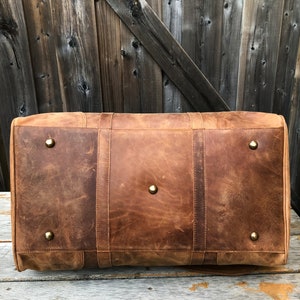K&ouml;nnte beinhalten: Eine braune Ledertasche mit Messing-Akzenten. Die Tasche hat eine rechteckige Form und ist aus abgenutztem Leder gefertigt.