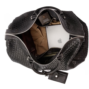 May include: Black woven leather travel bag with a zipper closure and a small leather pouch attached to the handle. The bag is open and contains a laptop, a brown shirt, and other items.