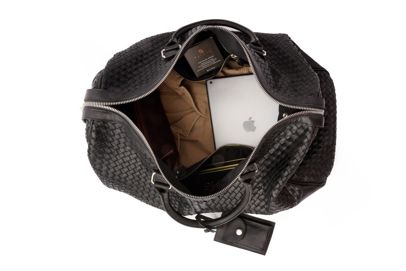 May include: Black woven leather travel bag with a zipper closure and a small leather pouch attached to the handle. The bag is open and contains a laptop, a brown shirt, and other items.