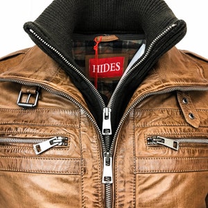 May include: A brown leather jacket with a zipper closure and multiple pockets. The jacket has a dark green knit collar and a red label with the word "HIDES" on it.
