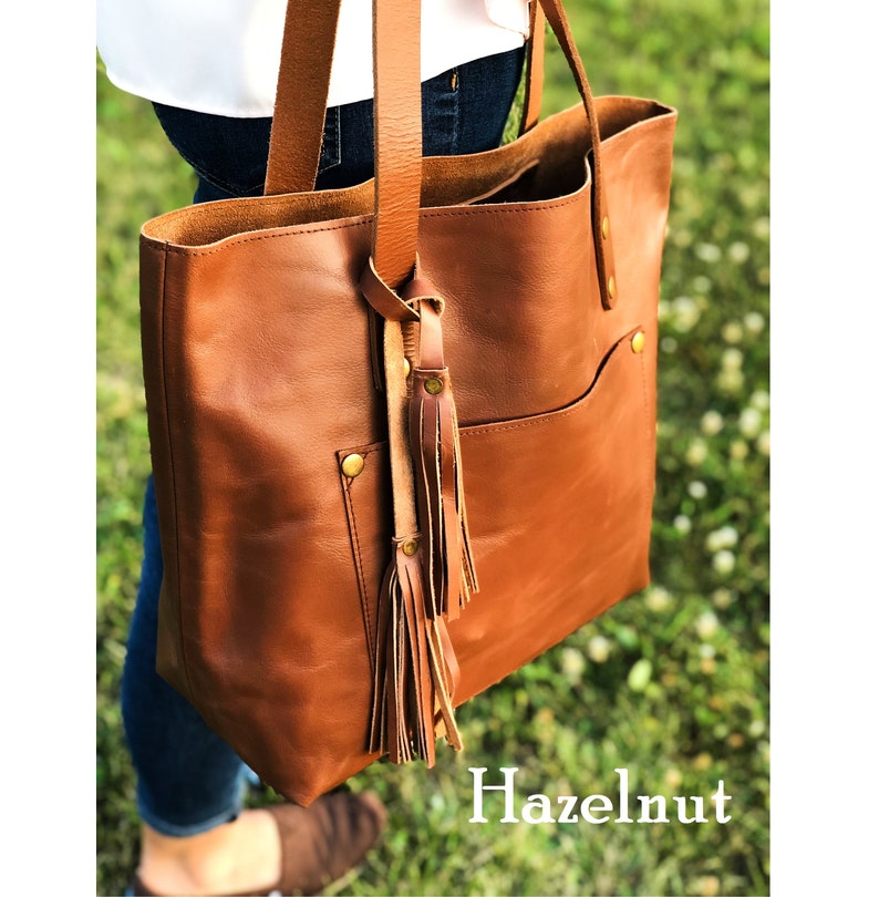 May include: A brown leather tote bag with a front pocket and a tassel. The bag has gold-coloured hardware and is labelled "Hazelnut".