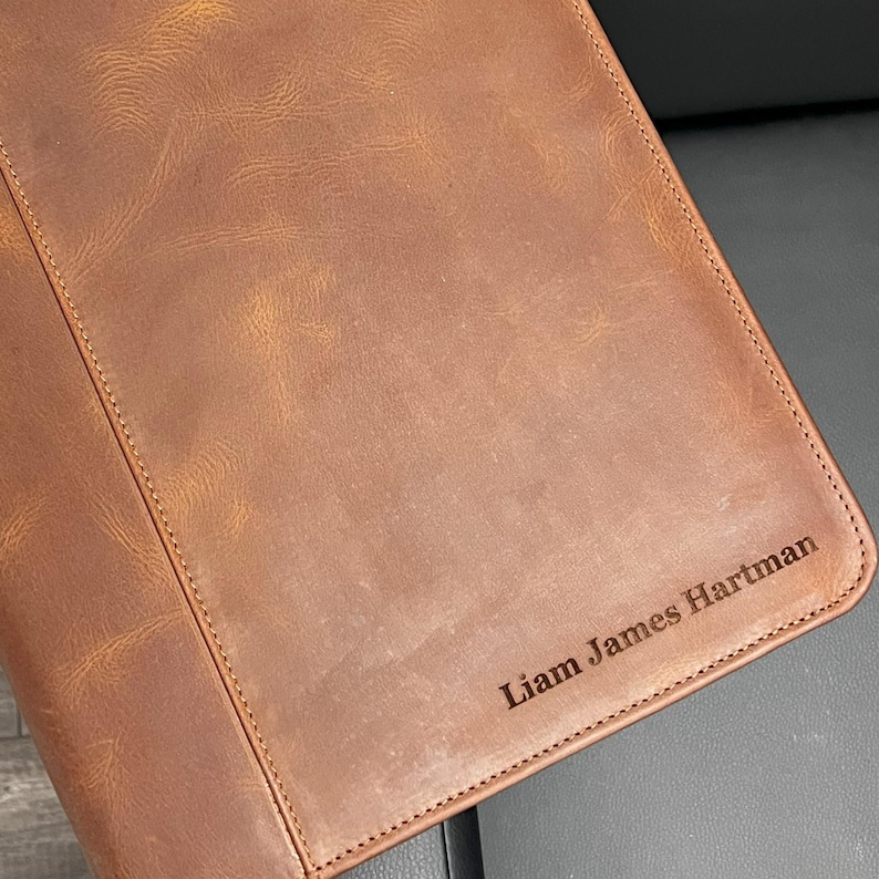 Może przedstawiać: Brązowy notes sk&oacute;rzany z wygrawerowanym imieniem "Liam James Hartman" na przedniej stronie.