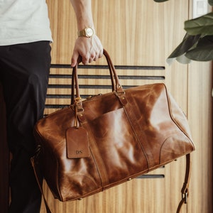 May include: A brown leather duffel bag with a shoulder strap and a leather tag that says "DS". The bag is sitting on a wooden floor. Saddle Brown.