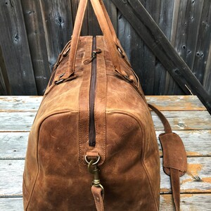 K&ouml;nnte beinhalten: Eine braune Ledertasche mit einem oberen Griff und einem Schultergurt. Die Tasche hat einen Rei&szlig;verschluss und einen Messingverschluss.