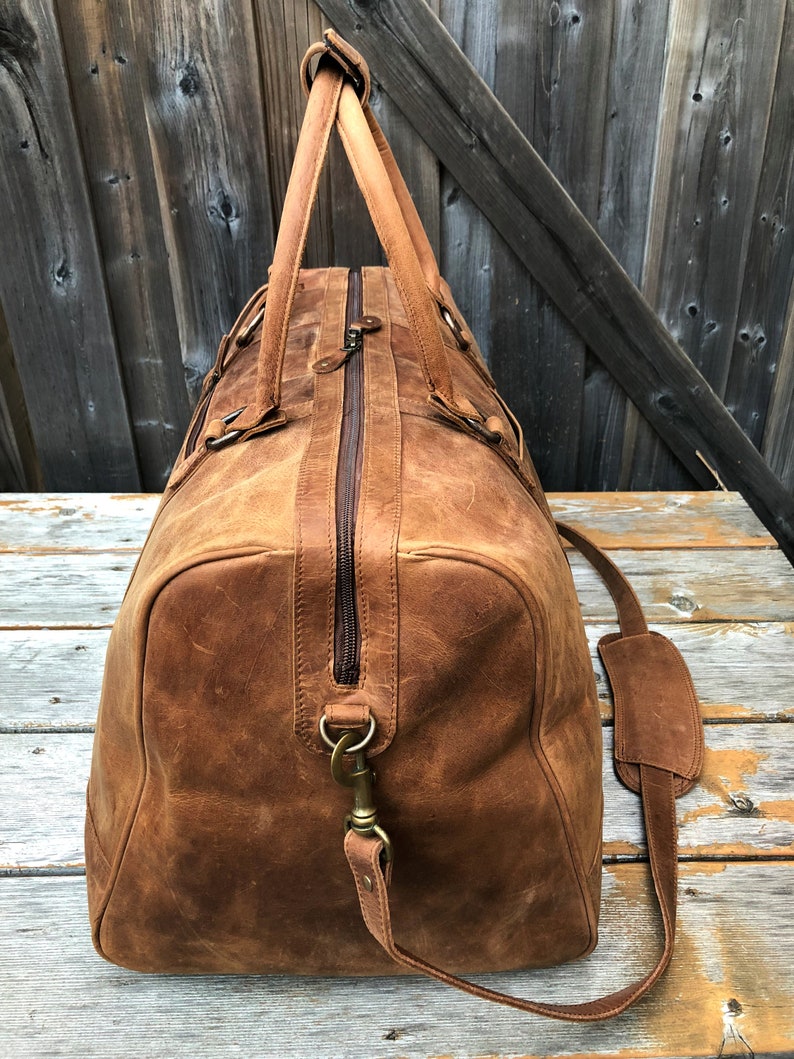 K&ouml;nnte beinhalten: Eine braune Ledertasche mit einem oberen Griff und einem Schultergurt. Die Tasche hat einen Rei&szlig;verschluss und einen Messingverschluss.