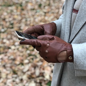 Puede incluir: Un par de guantes de conducir de cuero marr&oacute;n con perforaciones y cierre a presi&oacute;n. Los guantes los lleva una persona que sostiene un tel&eacute;fono inteligente.