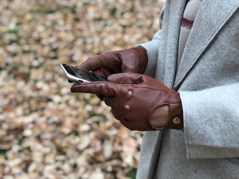 Puede incluir: Un par de guantes de conducir de cuero marr&oacute;n con perforaciones y cierre a presi&oacute;n. Los guantes los lleva una persona que sostiene un tel&eacute;fono inteligente.